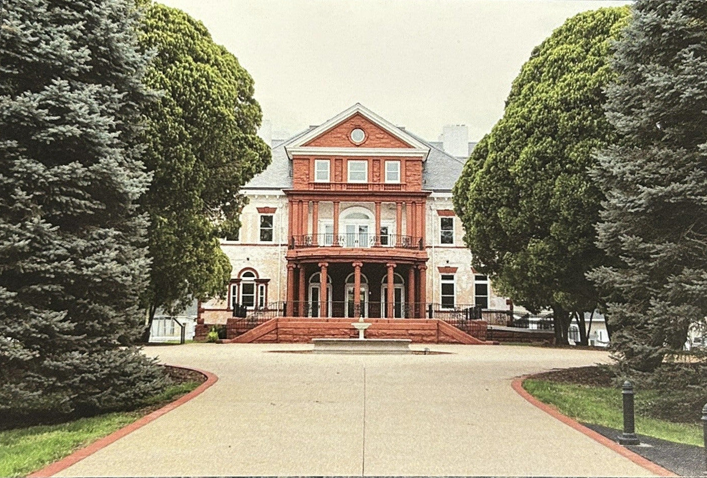 A rectangular art print or postcard showing a grand red brick mansion surrounded by tall green trees and a paved driveway.