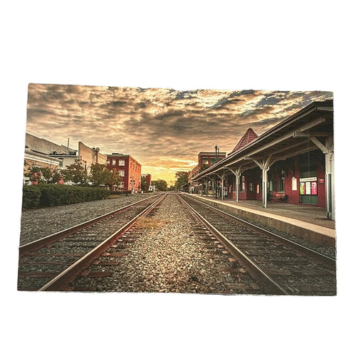 A rectangular art print depicting a scenic train station platform with two tracks under a dramatic sunset sky