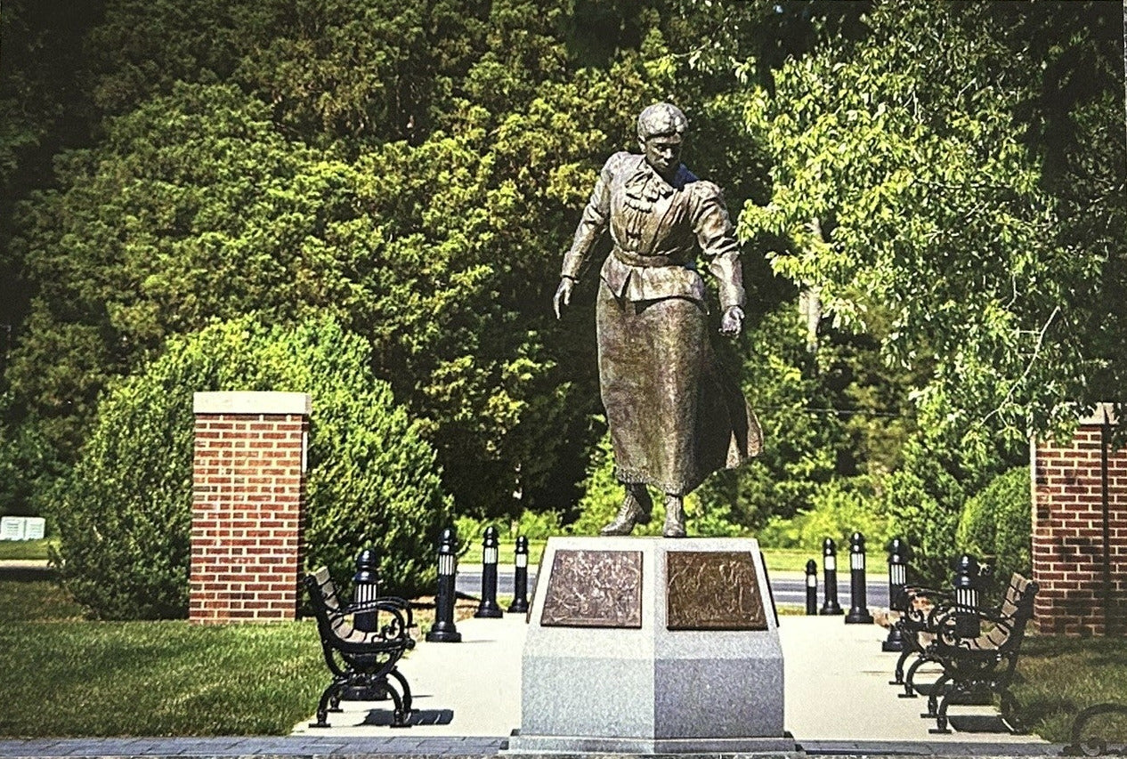 A rectangular printed photograph or postcard showing a bronze statue in a park-like setting with green trees, brick pillars, benches, and a stone pedestal, placed on a mottled gray stone surface.