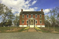 A rectangular printed photograph or postcard showing a red brick house with white trim, set in a grassy yard with trees and a blue sky