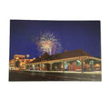 A rectangular printed photograph or postcard showing a nighttime train station scene with fireworks in the sky