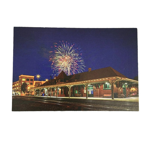 A rectangular printed photograph or postcard showing a nighttime train station scene with fireworks in the sky